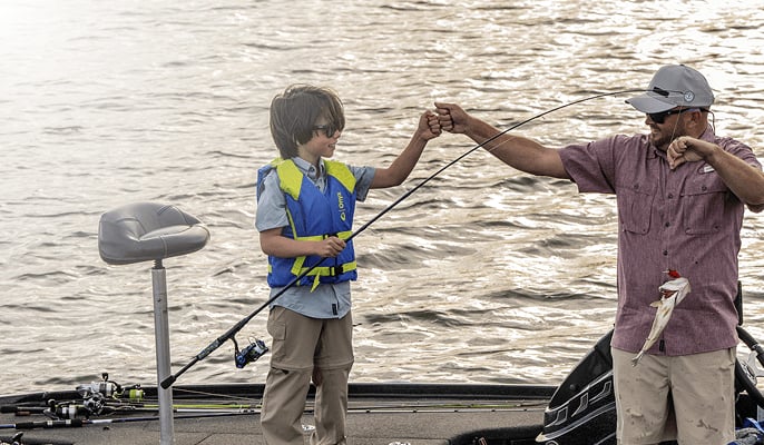  An adult and a child standing on a boat holding a fishing rod together as a fish is lifted from the water, with rippling water in the background.