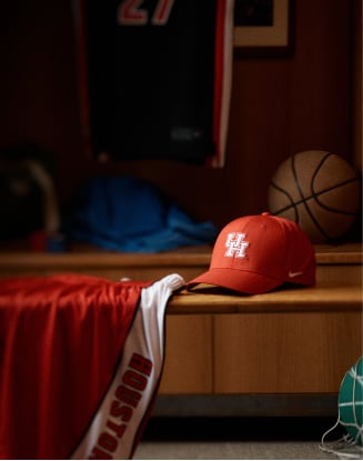 Red University of Houston baseball-style cap with white embroidered ‘UH’ logo resting on a wooden locker bench, next to a red-and-white HOUS– branded athletic jersey and a basketball in the background.